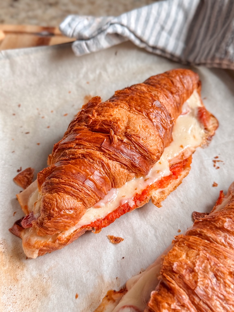 Golden brown pizza croissant sandwich on a parchment-lined baking sheet with melted mozzarella and marinara sauce.